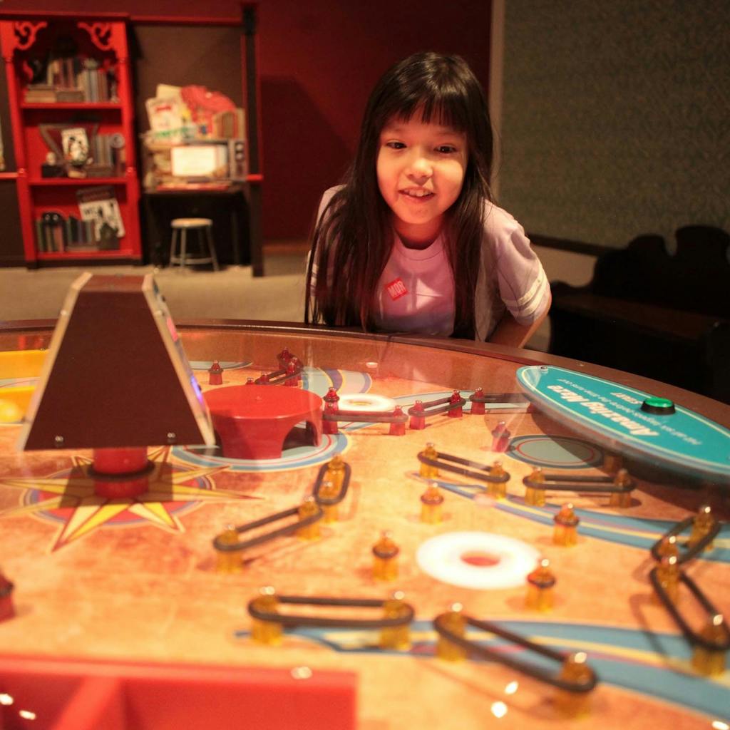 A young girl looks in front of her at a large circular table with small balls rolling through a colorful maze.